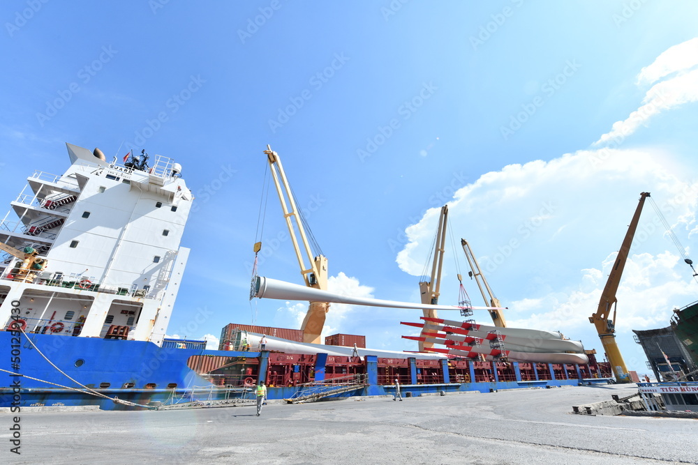 Obraz premium Large wind turbine rotor blades being unloaded from a ship onto a telescopic trailer for onward transportation by road to the construction site