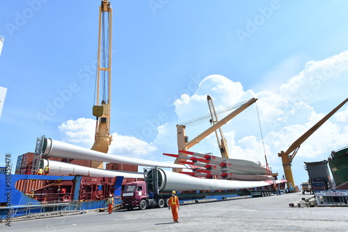 Large wind turbine rotor blades being unloaded from a ship onto a telescopic trailer for onward transportation by road to the construction site