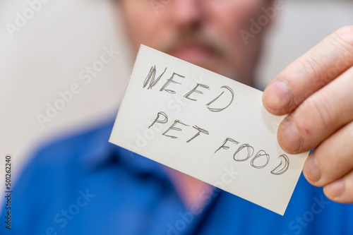 Need pet food. It is written in jagged gray letters on the paper. Man holding a white paper rectangle with handwritten text in front of his face. Close-up of a mature man's hand.