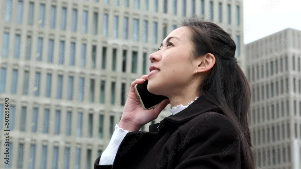 Low angle side view of happy Asian businesswoman in smart casual clothes. She is smiling and looking up while speaking on cellphone against skyscrapers on city street Low angle side view of happy Asian businesswoman in smart casual clothes. She is smiling and looking up while speaking on cellphone against skyscrapers on city street