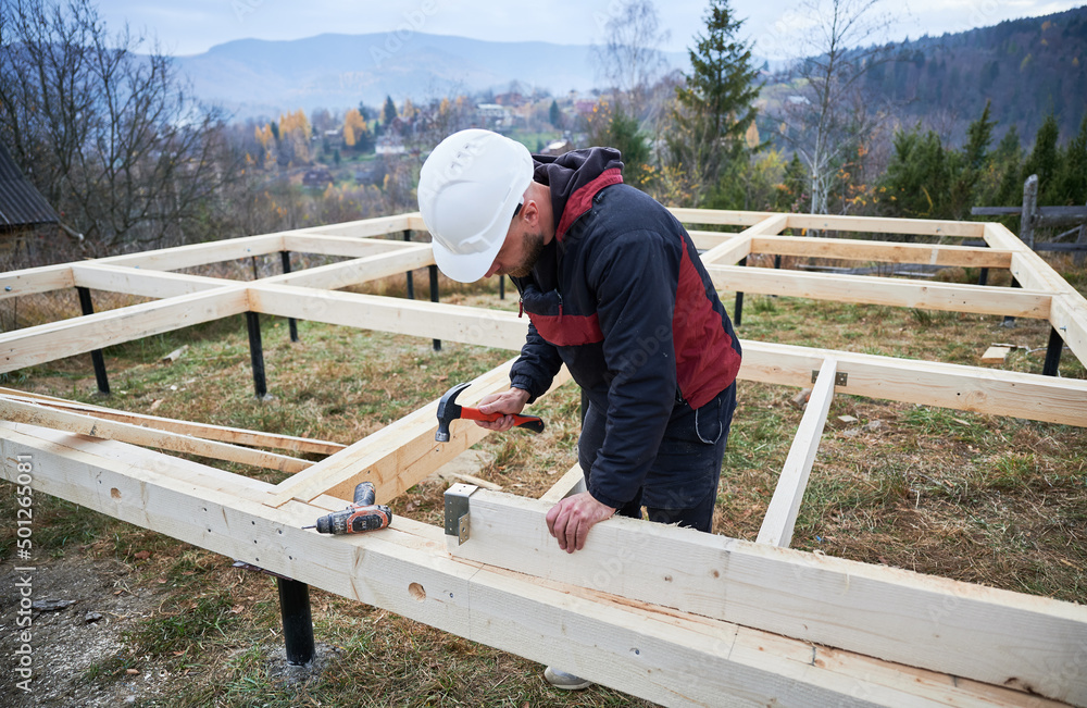 Man worker building wooden frame house on pile foundation. Carpenter installing furniture for wooden joist, using hammer and screwdriver. Carpentry concept. Man worker building wooden frame house on pile foundation. Carpenter installing furniture for wooden joist, using hammer and screwdriver. Carpentry concept.