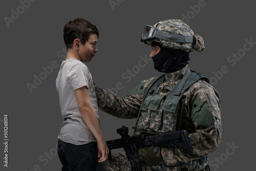 Shot of brave soldier with rifle protecting little child isolated on gray background.