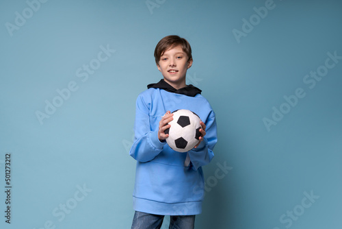 Smiling teen boy in sports blue clothes in a sweatshirt holding soccer ball posing in studio on a colored background. Caucasian teenager fan of playing football