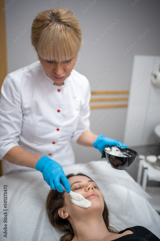 Doctor cosmetologist or dermatologist making face mask in cosmetology salon. Professional Beautician applying face mask on caucasian woman face lying on bed in bathrobe. Doctor cosmetologist or dermatologist making face mask in cosmetology salon. Professional Beautician applying face mask on caucasian woman face lying on bed in bathrobe.