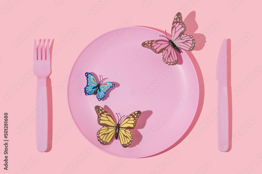 Spring creative layout with pastel pink plate, fork,knife and colorful butterfly on pastel pink background. 80s or 90s aesthetic fashion food restaurant concept. Minimal romantic summer idea.