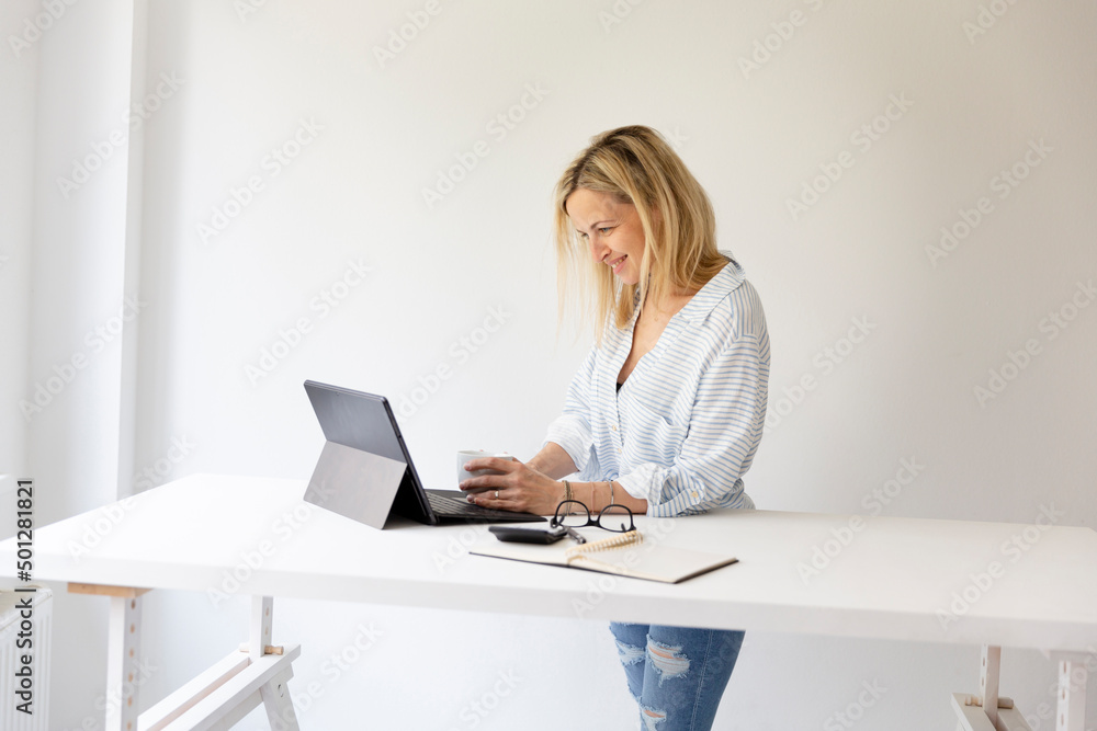 pretty young blonde business woman stands by her ergonomic height adjustable work desk and works on tablet from home in her home office pretty young blonde business woman stands by her ergonomic height adjustable work desk and works on tablet from home in her home office
