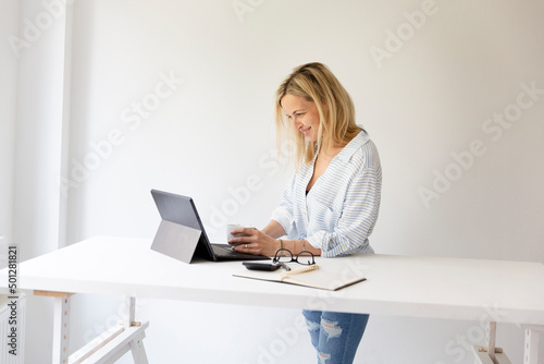 pretty young blonde business woman stands by her ergonomic height adjustable work desk and works on tablet from home in her home office