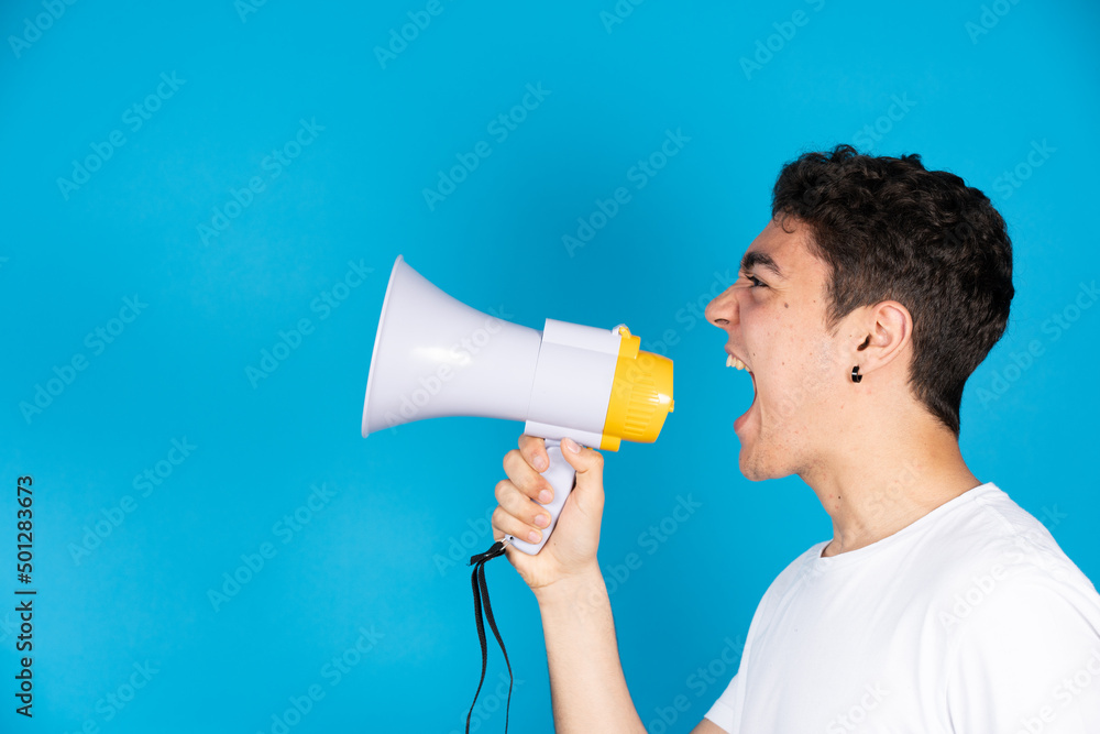 Hispanic teenager boy shouting on megaphone or speaker to copy space isolated on blue background. Hispanic teenager boy shouting on megaphone or speaker to copy space isolated on blue background.