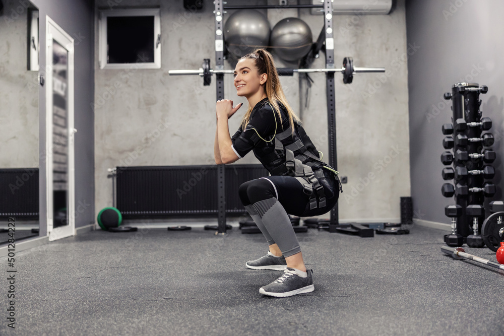 Fototapeta premium A beautiful woman in an EMS suit does squats in a gym.
