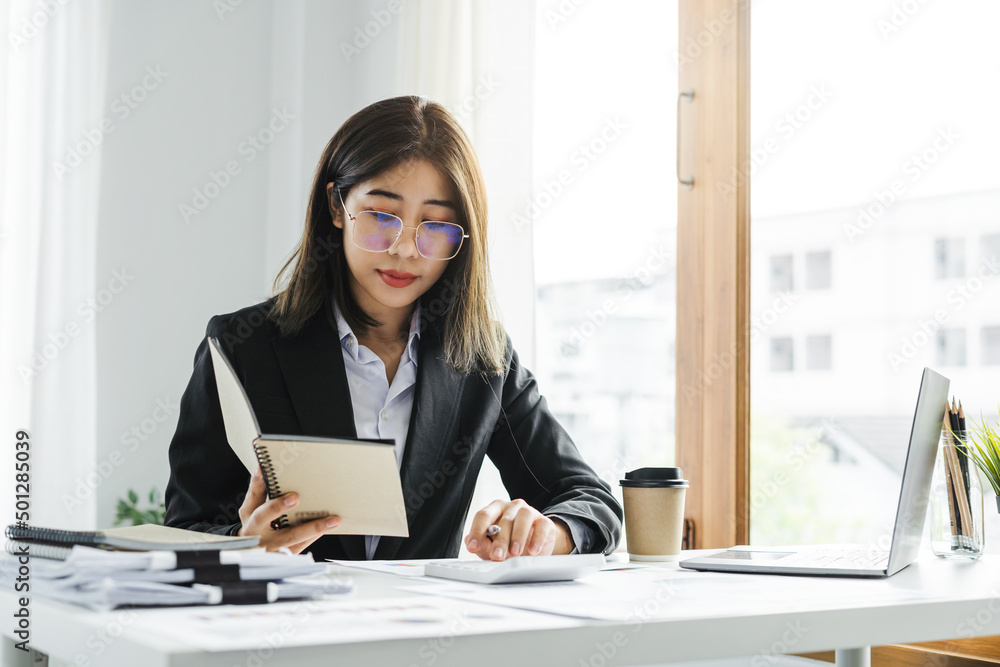Portrait of a short-haired Asian businesswoman using a notebook with a calculator to calculate accounting taxes in a modern office.