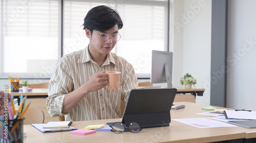 Handsome male office worker holding coffee cup and using computer tablet on wooden desk.