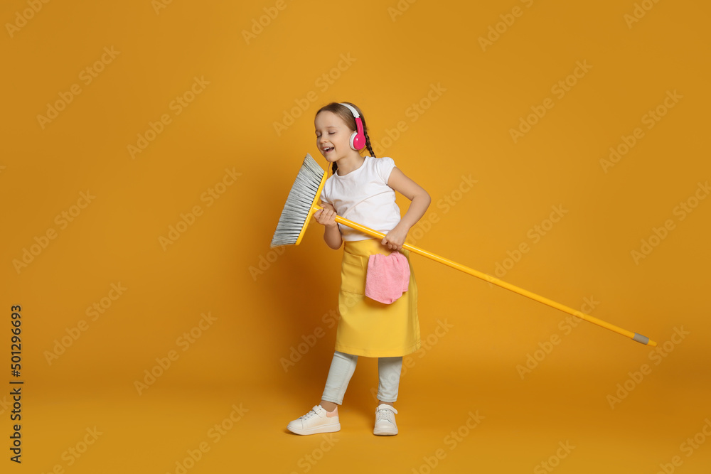 Cute little girl in headphones with broom singing on orange background Cute little girl in headphones with broom singing on orange background