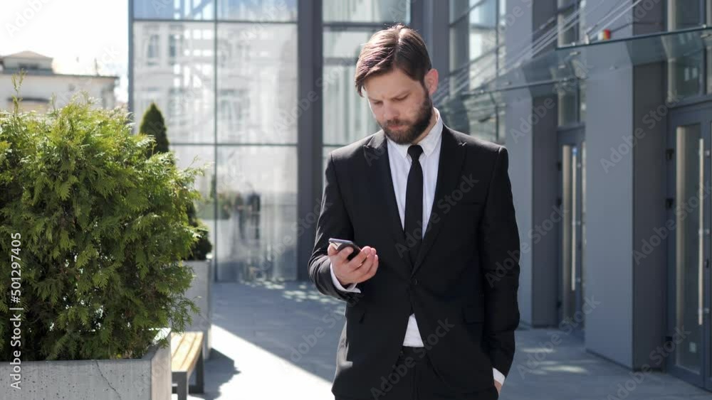 Portrait of adult businessman takes a cell phone out of his pocket reading sms texting message checking email while smiling. Successful male business man standing near modern glass office outdoor. Portrait of adult businessman takes a cell phone out of his pocket reading sms texting message checking email while smiling. Successful male business man standing near modern glass office outdoor.