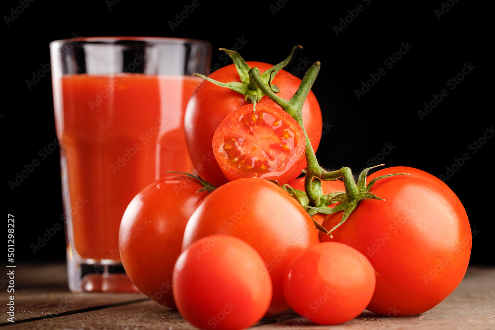 A glass cup with tomato juice, next to it lies a branch of tomatoes. On a wooden table there is a glass of tomato juice and tomatoes lie on a black background. A glass cup with tomato juice, next to it lies a branch of tomatoes. On a wooden table there is a glass of tomato juice and tomatoes lie on a black background.