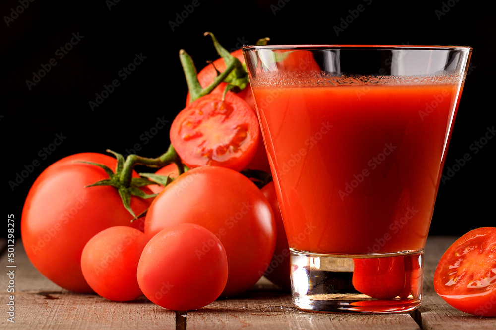 A glass cup with tomato juice, next to it lies a branch of tomatoes. On a wooden table there is a glass of tomato juice and tomatoes lie on a black background. A glass cup with tomato juice, next to it lies a branch of tomatoes. On a wooden table there is a glass of tomato juice and tomatoes lie on a black background.