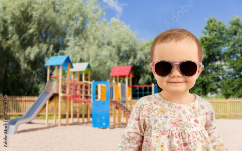 childhood, summer and people concept - happy little baby girl in sunglasses over children's playground background