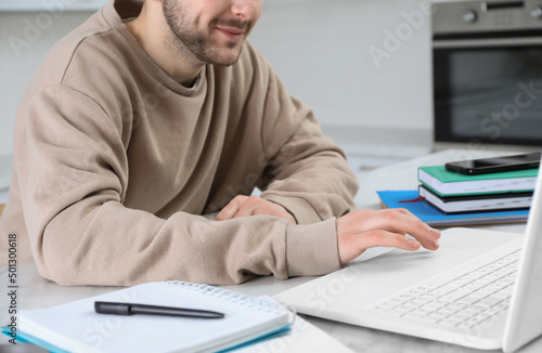 Young man using modern laptop for studying in kitchen, closeup. Distance learning