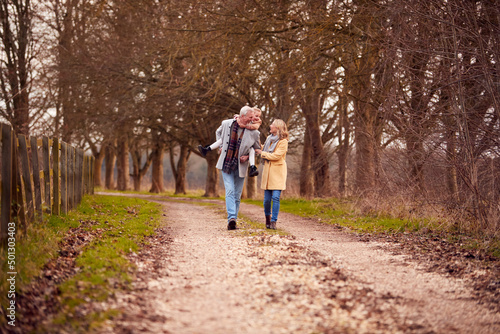 Grandfather Gives Granddaughter Piggyback Walking Through Winter Countryside With Grandmother