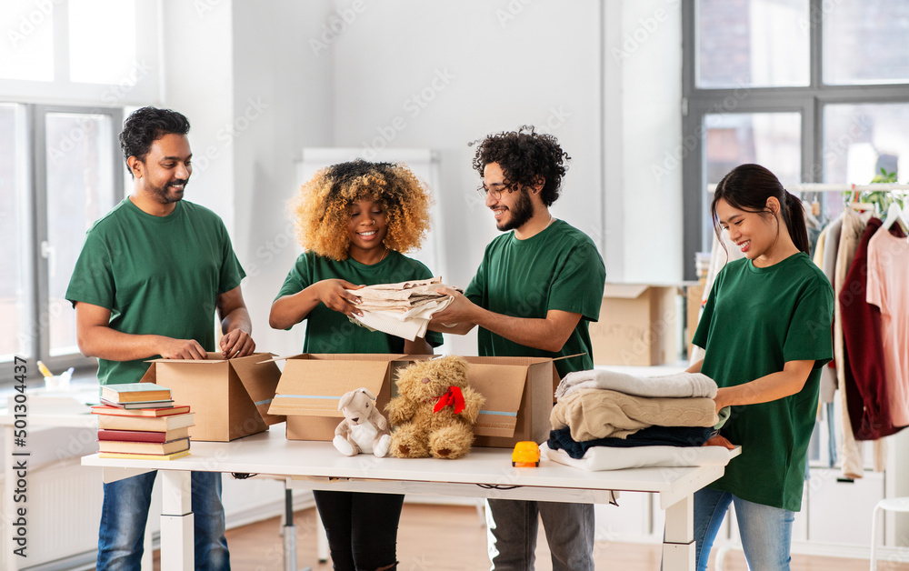 Obraz premium charity, donation and volunteering concept - international group of happy smiling volunteers packing clothes and other stuff in boxes at distribution or refugee assistance center