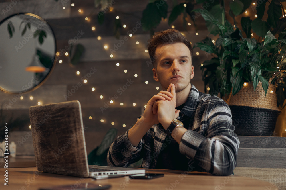 Pensive young man sitting at table with laptop and thinking about project. Male freelancer thoughtfully looking away during remote work on online startup plan in cafe Pensive young man sitting at table with laptop and thinking about project. Male freelancer thoughtfully looking away during remote work on online startup plan in cafe