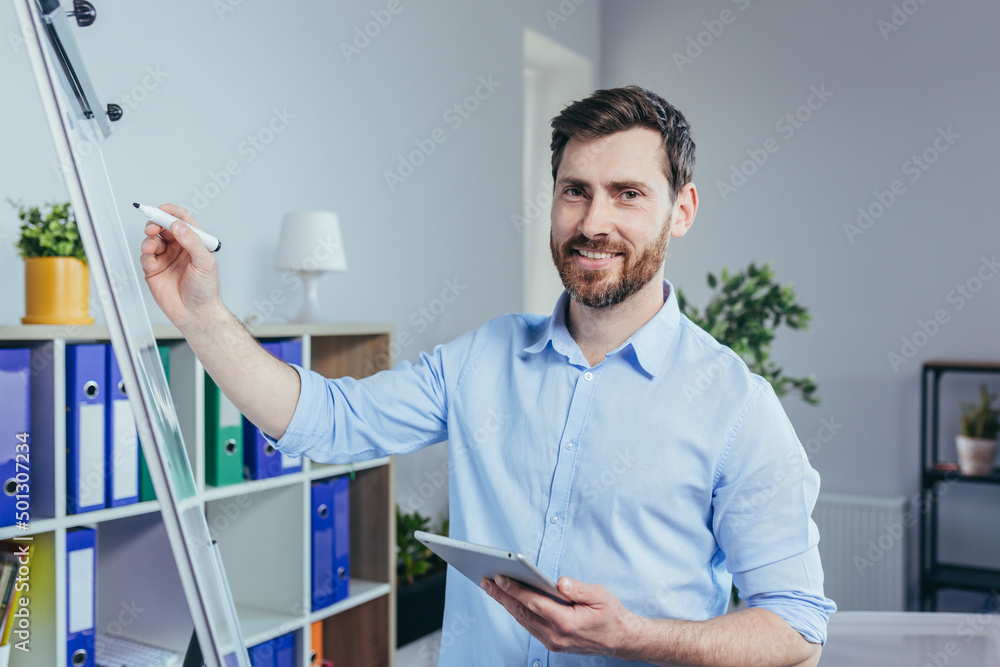 Portrait of a successful business founder, a man looks at the camera and smiles at a white board for notes, holding a tablet, describes a business strategy Portrait of a successful business founder, a man looks at the camera and smiles at a white board for notes, holding a tablet, describes a business strategy