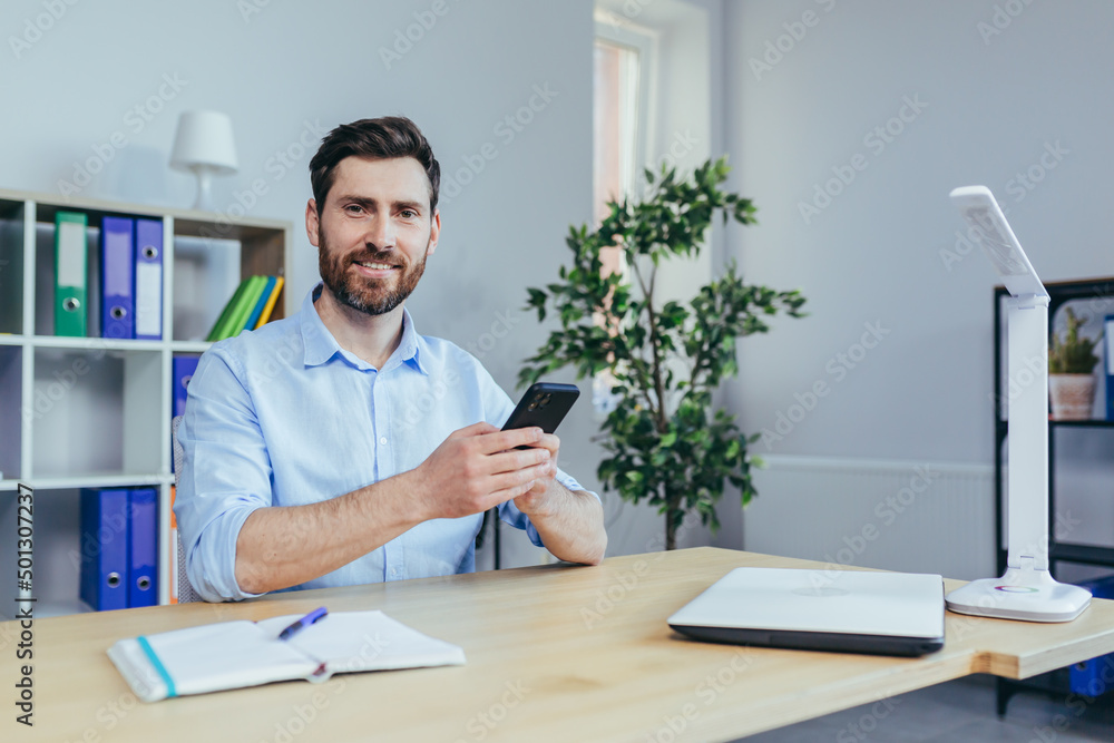 Portrait of a successful businessman in the office, a man sitting at a table and holding a phone, happy smiling Portrait of a successful businessman in the office, a man sitting at a table and holding a phone, happy smiling