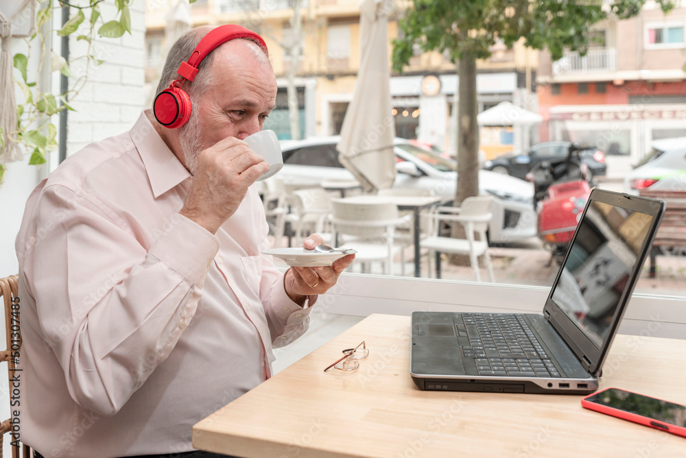 smiling older man with headphones enjoying his cup of coffee, using laptop for language learning, online classes, enjoying his retirement smiling older man with headphones enjoying his cup of coffee, using laptop for language learning, online classes, enjoying his retirement