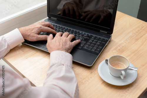 the hands of an unrecognizable older man typing on the keyboard of a laptop, a cup of coffee on the table
