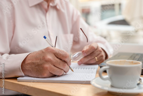 close-up of the hands of an unrecognizable older man making notes in his notebook, holding his glasses in one hand, a cup of coffee next to him