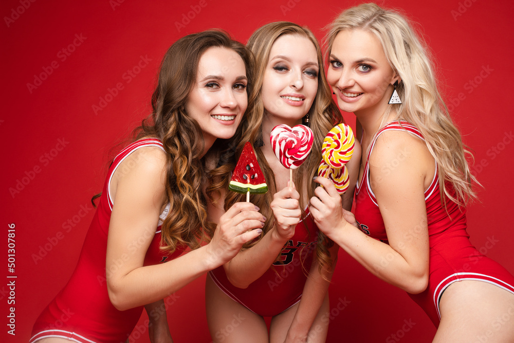 Beautiful and happy female friends in bright red swimsuits with bright candy lollipops in their hands on a red background, studio.
