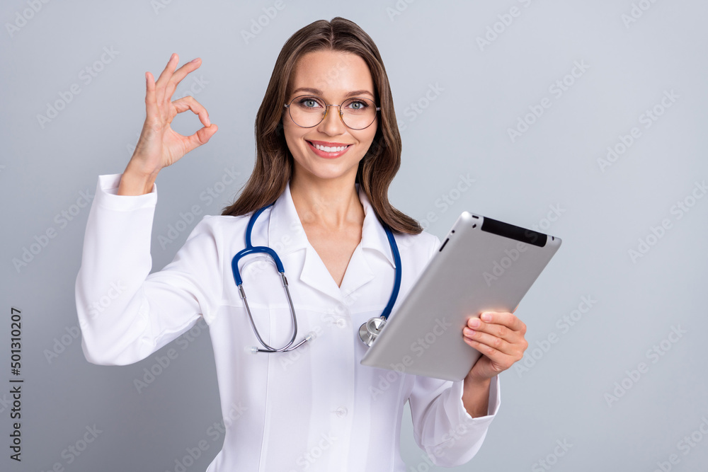Photo of young woman cardiologist use modern device show fingers okey symbol recommend isolated over grey color background Photo of young woman cardiologist use modern device show fingers okey symbol recommend isolated over grey color background