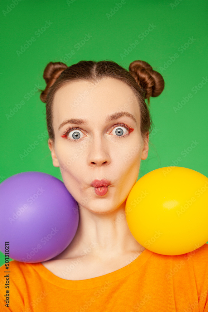 Young woman making face with purple and yellow balloons against green background Young woman making face with purple and yellow balloons against green background