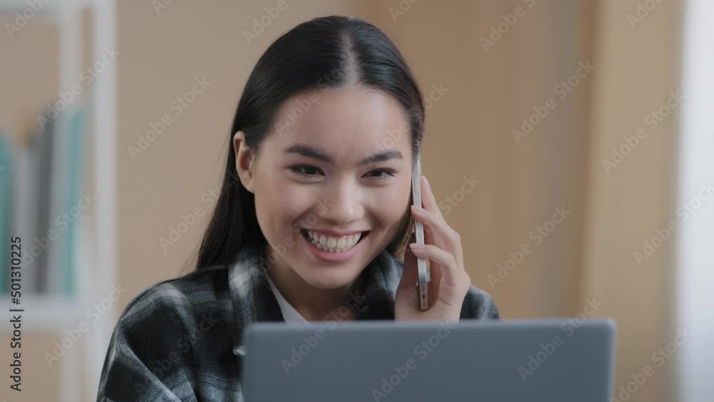 Happy smiling Asian girl Korean female freelancer chatting with friends on phone answering call friendly looking at laptop screen remote working business negotiation making online order by smartphone Happy smiling Asian girl Korean female freelancer chatting with friends on phone answering call friendly looking at laptop screen remote working business negotiation making online order by smartphone