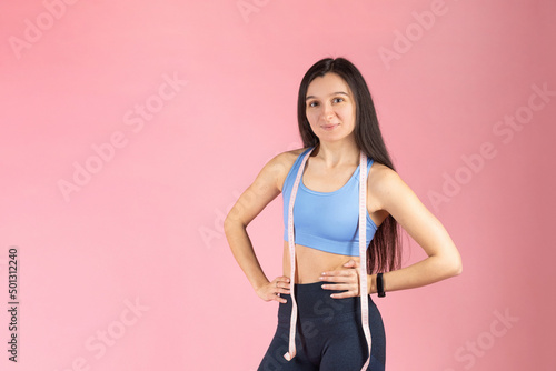 The trainer holds in his hands a measuring tape to measure the body. Attractive brunette woman in fashionable sportswear on pink background. Healthy lifestyle concept © mdyn