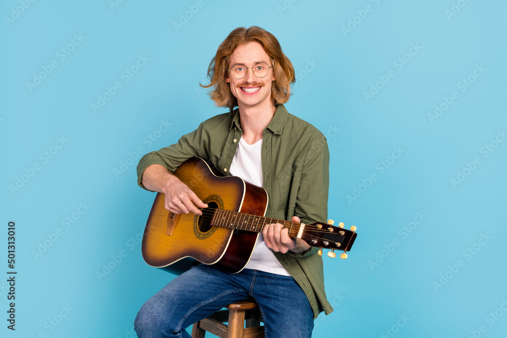 Portrait of attractive cheerful talented guy sitting on stool playing guitar live hit isolated over pastel blue color background Portrait of attractive cheerful talented guy sitting on stool playing guitar live hit isolated over pastel blue color background