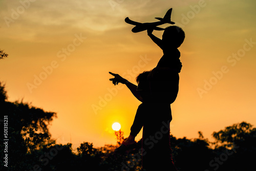 boy holding toy plane and sitting on his father shoulders and having fun together outdoors twilight, silhouette image.