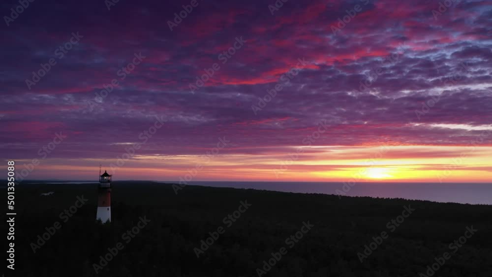 Beautiful sunset over green forest. View of the lighthouse, sea in the background. Orange sun, pastel pink clouds, red sky. Beautiful sunset over green forest. View of the lighthouse, sea in the background. Orange sun, pastel pink clouds, red sky.