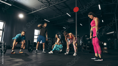 Group of young friends doing sports, training at gym indoors, lifiting weights, dumbbells