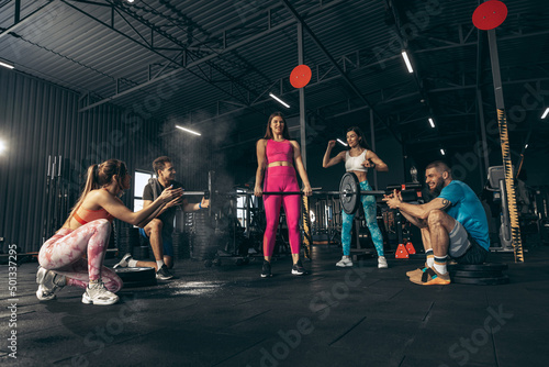 Group of young friends doing sports, training at gym indoors. Girl in bright sportswear lifting heavy barbell