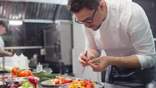 Restaurant chef in uniform and glasses adding fresh dill and pickled onion to tomato salad on plate while garnishing dish before serving photo