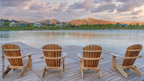 Panorama Puffy clouds at sunset Four wooden lounge chairs facing the reflective Oquirrh Lake at © Jason