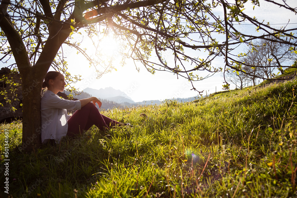 young brunette girl looking at infinity leaning against the trunk of a tree at sunset. rural area, grassy field with mountains in the background. young brunette girl looking at infinity leaning against the trunk of a tree at sunset. rural area, grassy field with mountains in the background.