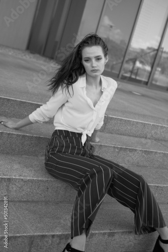 A young brunette girl in white shirt and striped trouser is sitting on the stairs