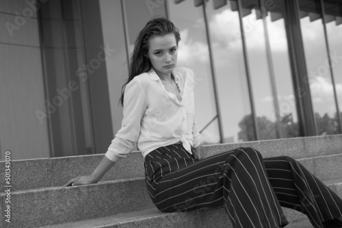 A young brunette girl in white shirt and striped trouser is sitting on the stairs