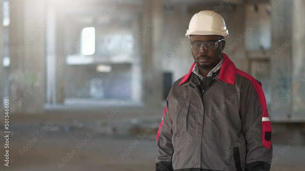 African american worker in white hard hat and protective goggles stands at construction area and looks at the camera African american worker in white hard hat and protective goggles stands at construction area and looks at the camera