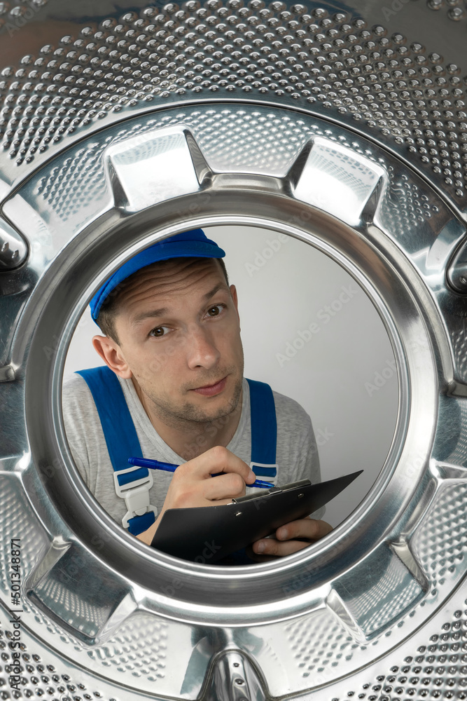 Male repairman in uniform with a tablet in his hands looks into the drum of a washing machine, photo from the inside. Vertical photo Male repairman in uniform with a tablet in his hands looks into the drum of a washing machine, photo from the inside. Vertical photo