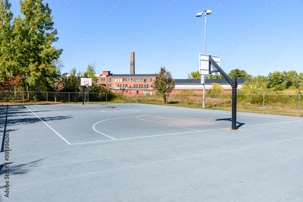 Empty basketball court in a public park on a clear autumn day. A old industrial building is visible in background.