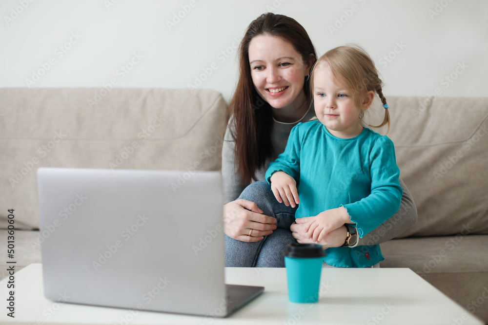 Fototapeta premium Mother and her daughter using video chat on laptop sitting on cozy sofa at home, watching video, having fun, two generations