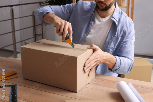 Man using utility knife to open parcel at wooden table indoors, closeup photo