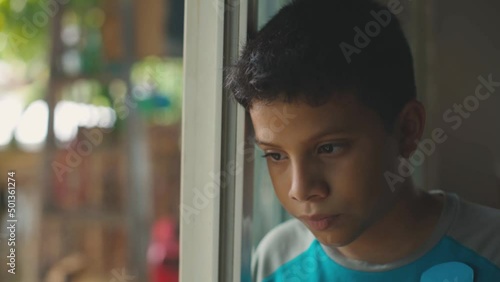 Hispanic young man leaning against the window, looking out, very thoughtful.